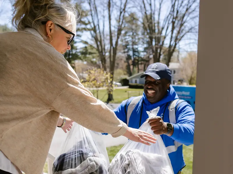Smiling driver in a blue uniform taking clear bags of laundry from a customer on a suburban lawn