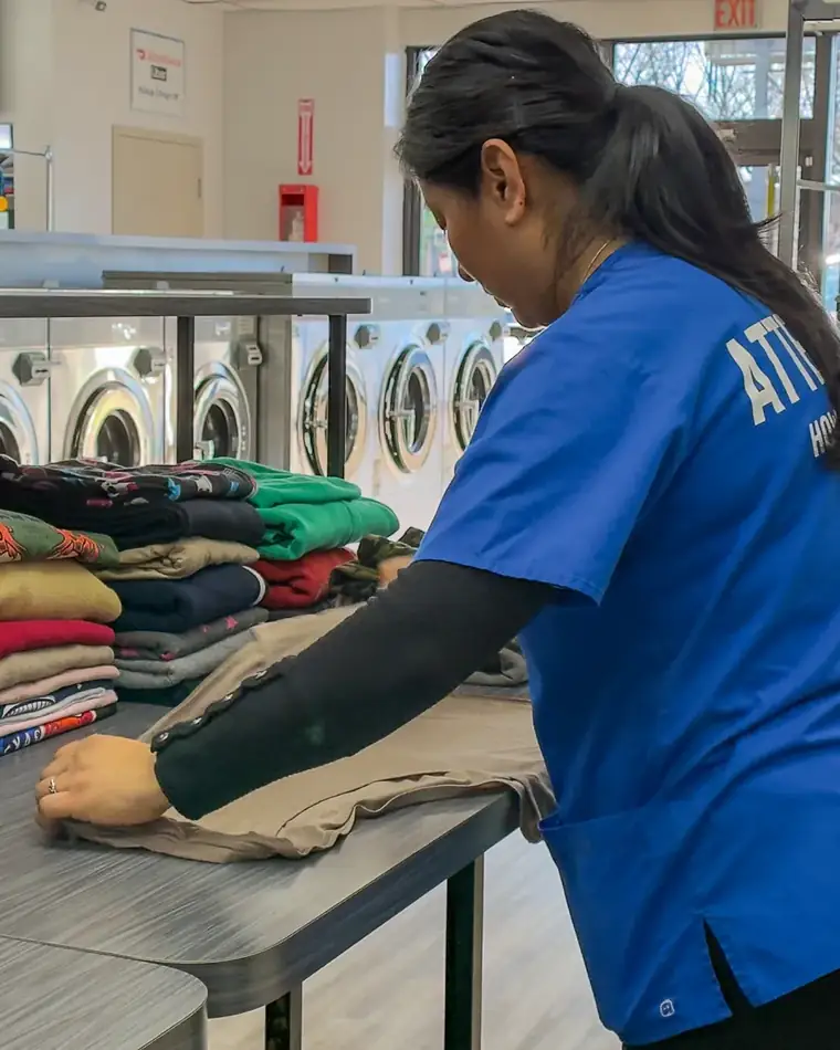 Attendant in a blue shirt folding a garment at a work table with stacks of folded clothes and washing machines behind