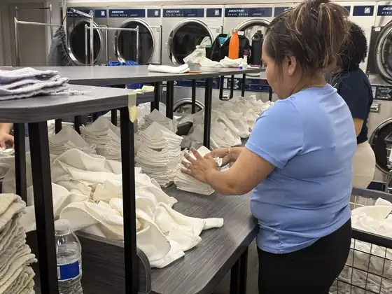 Employee in a light blue shirt folding white hotel towels at a table in a commercial laundry room