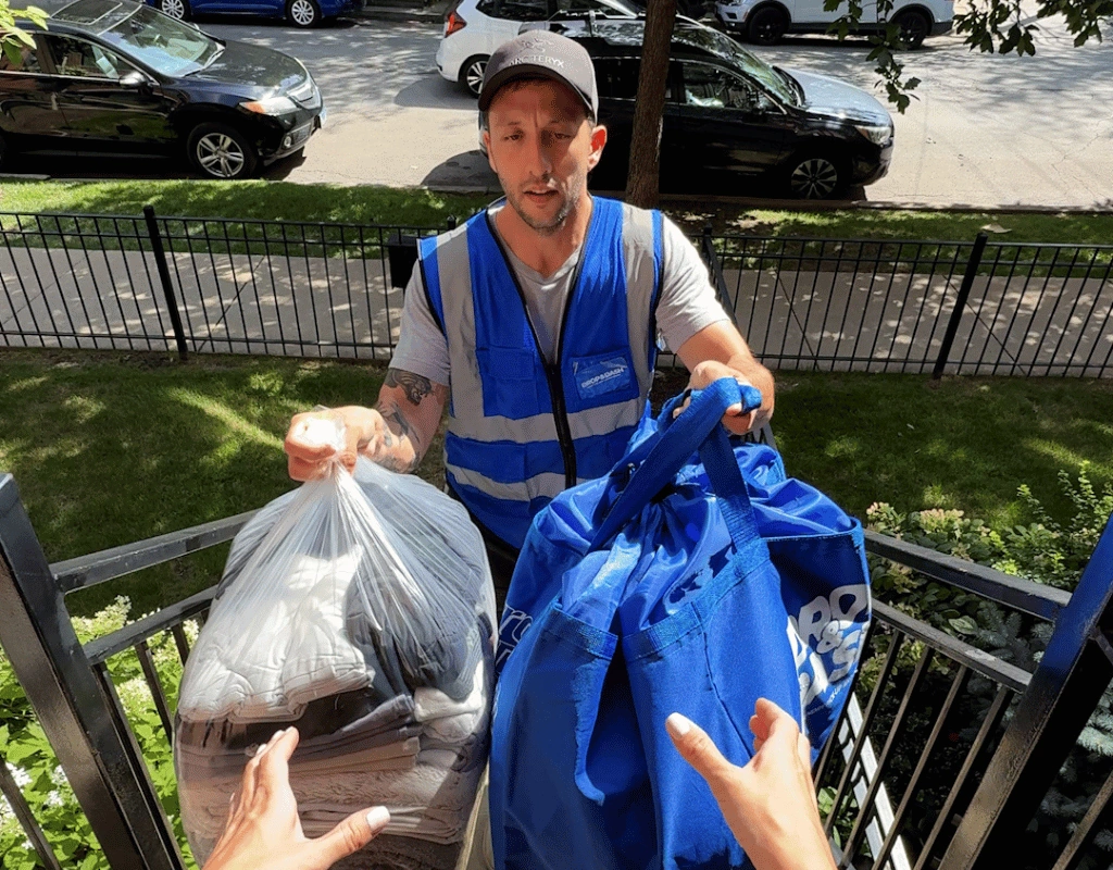 Driver in a reflective safety vest passing a clear bag and blue totes of laundry to a customer outdoors
