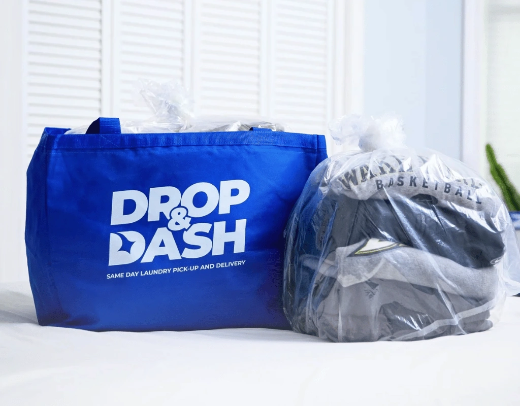 Blue Drop & Dash tote bag next to a clear sealed bag of folded clothing on a white bed