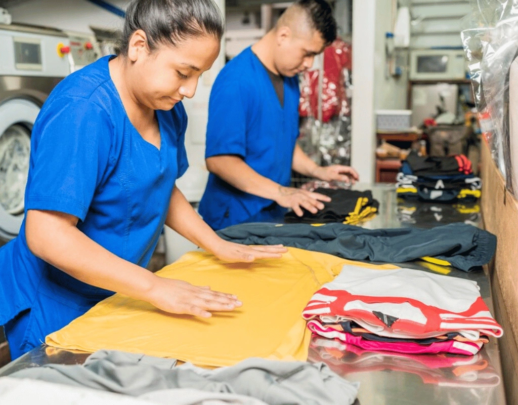 Two employees in blue shirts folding brightly colored shirts at a work table in a laundry facility