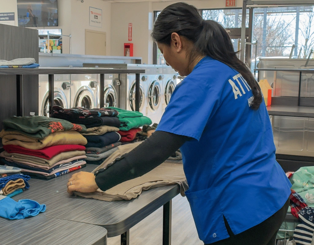 Attendant in a blue shirt folding stacks of towels and linens at a long table in front of commercial washers