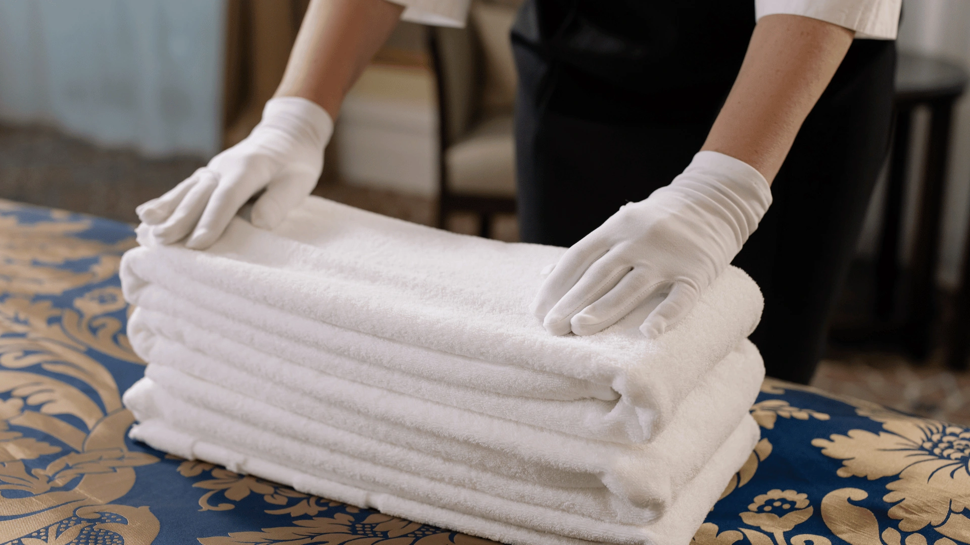 Gloved hands setting a stack of folded white towels on a patterned blue hotel bedspread