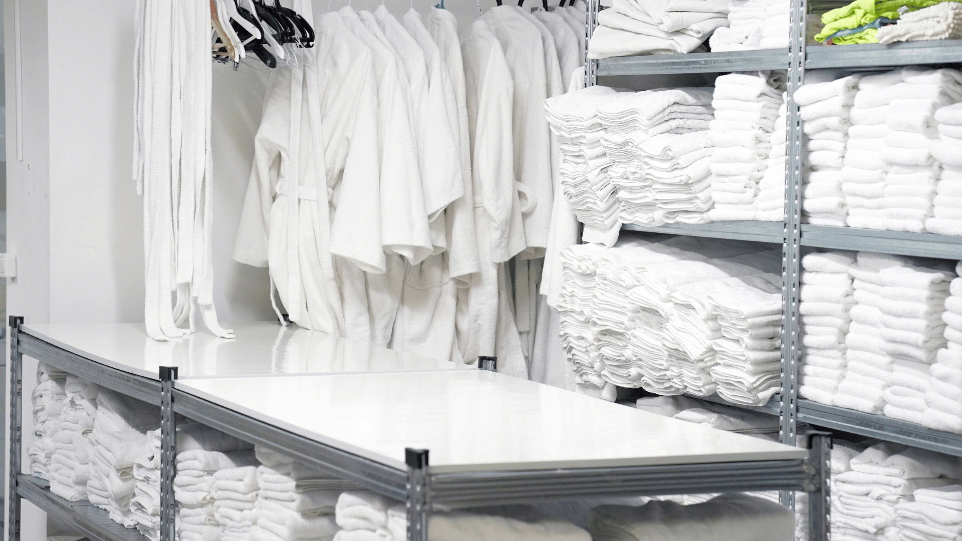 Rack of white medical coats on hangers beside metal shelves stacked with folded white towels and linens