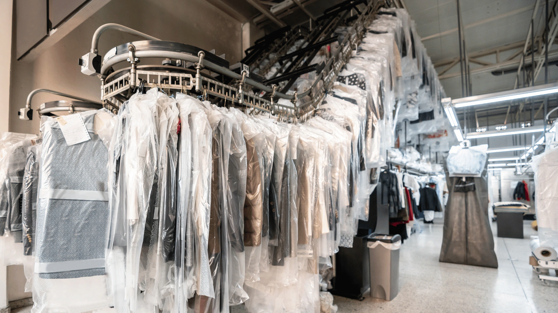 Rows of plastic-covered garments hanging on an overhead conveyor rail inside a large cleaning plant