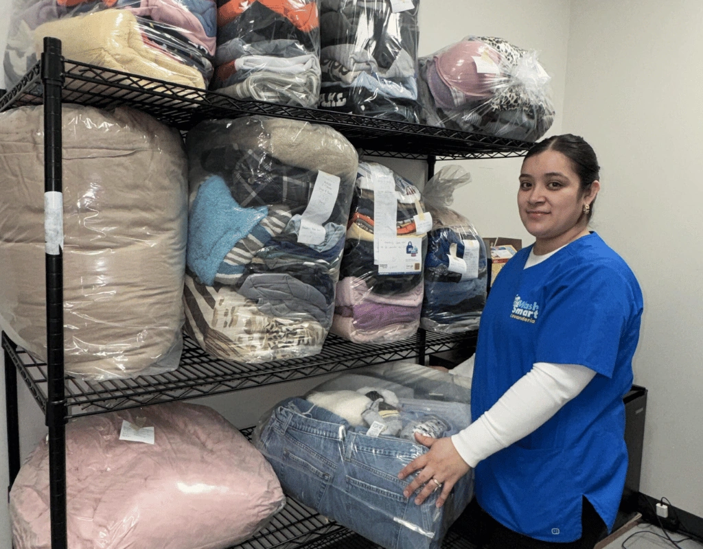 Employee in a blue shirt standing beside black wire shelves holding wrapped bundles of blankets, pillows and folded clothing