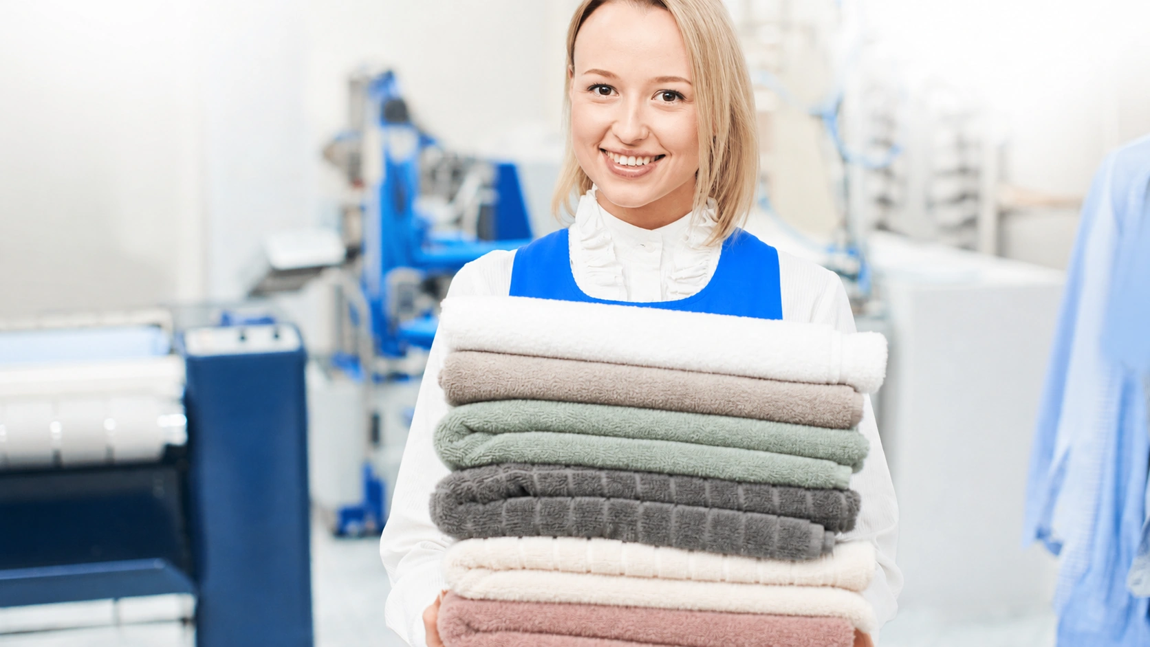 Smiling employee in a blue apron holding a stack of folded towels in a laundry facility