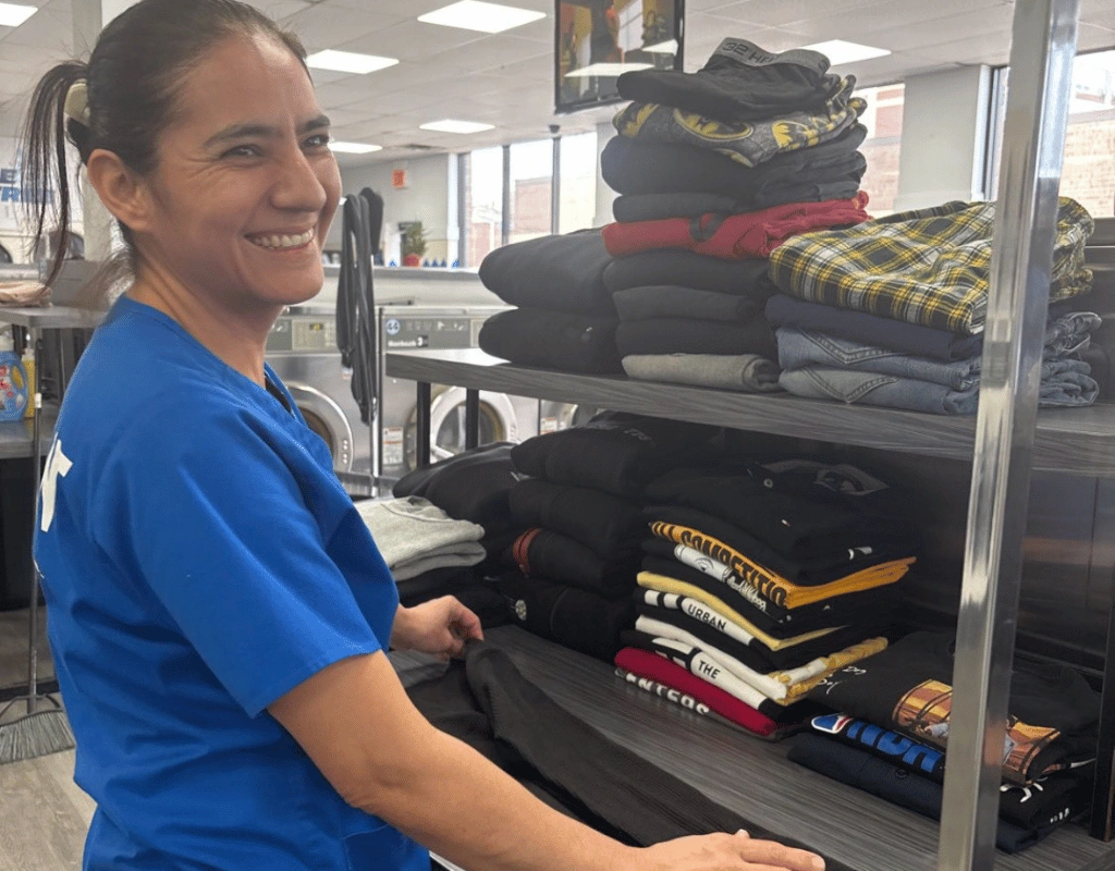 Smiling attendant in a blue shirt beside shelves of folded dark uniforms, plaid shirts and towels