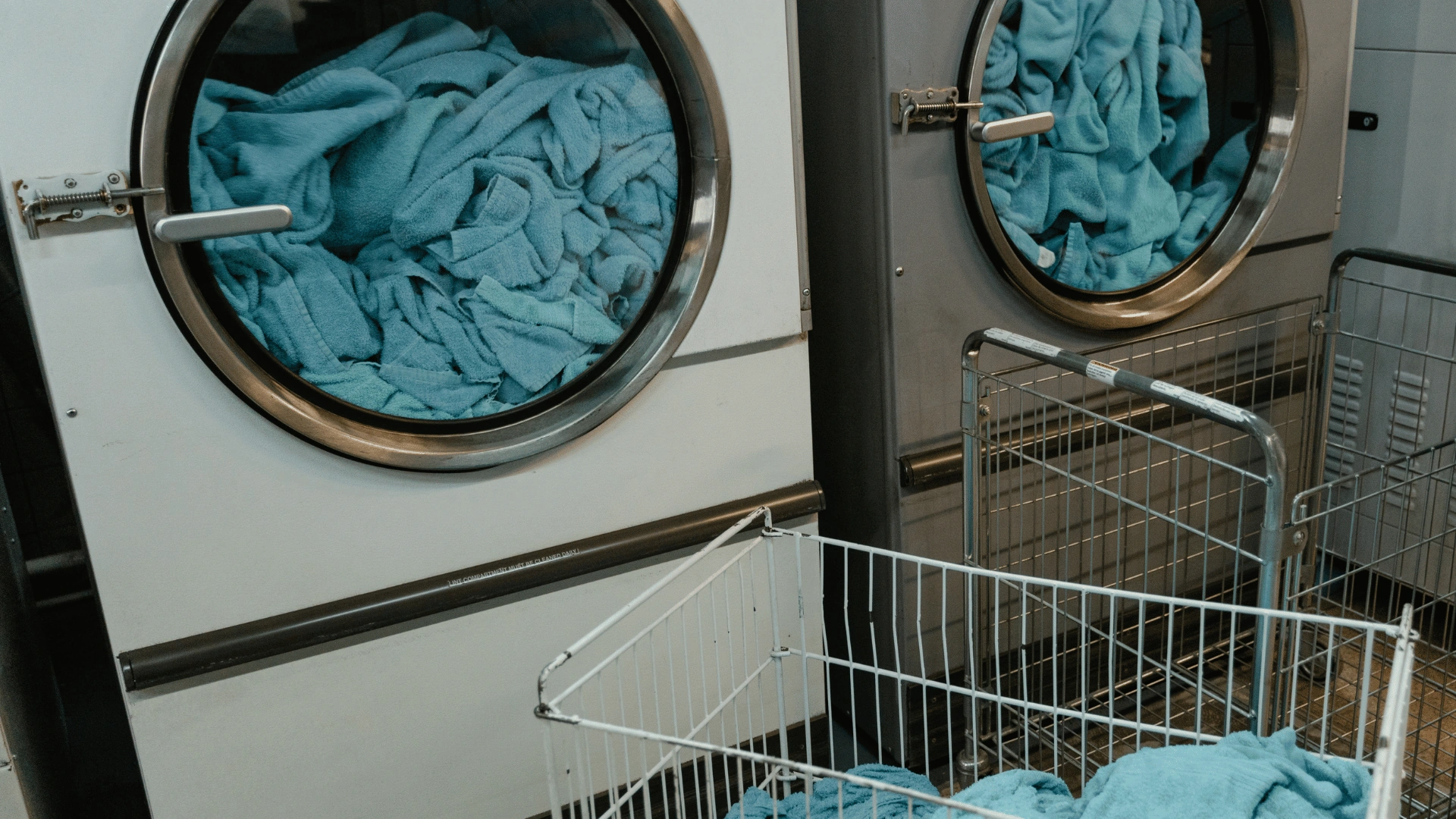 Two commercial washing machines loaded with teal gym towels beside an empty wire laundry cart