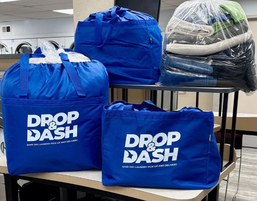 Three blue Drop & Dash tote bags and wrapped laundry bundles arranged on a table in a laundry facility