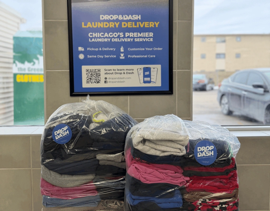 Sealed bundles of folded laundry on a window ledge below a framed Drop & Dash laundry delivery poster